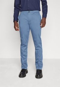 Pantaloni in cotone azzurro chiaro con vestibilità slim, caratterizzati da chiusura con bottone e tasche. Indossati con scarpe nere su uno sfondo neutro.