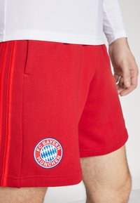Shorts de sport rouges en tissu léger, avec un logo FC Bayern München bleu et blanc et des poches latérales.