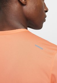 Camiseta atlética naranja con cuello redondo y mangas cortas, con una franja de acento gris en la parte posterior cerca del cuello.
