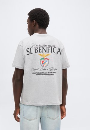 Άνδρας που φοράει ανοιχτό γκρι μπλουζάκι SL BENFICA με το λογότυπο της ομάδας και κείμενο στην πλάτη, συνδυασμένο με ανοιχτό μπλε τζιν, στέκεται μπροστά σε λευκό φόντο.
