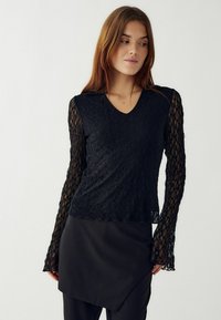 Čierny čipkový top s výstrihom do V, dlhými priesvitnými rukávmi a mierne rozšíreným spodným lemom. Obsahuje zložité kvetinové vzory po celej ploche.
