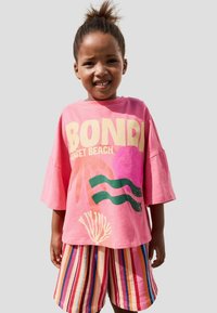 Cămașă grafică roz cu textul "BONDI SUNSET BEACH" și imprimeuri colorate. Asortată cu șorturi striped în nuanțe de roz, roșu, albastru și galben.