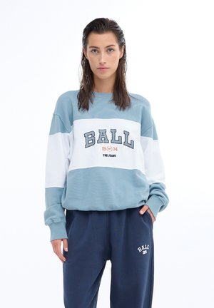 Felpa oversize azzurro chiaro e bianca con stampa "BALL"; scollatura a girocollo, maniche lunghe, texture morbida. Abbinata a pantaloni della tuta blu scuro.