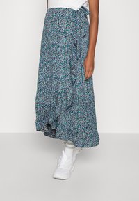 Květinová zavinovací sukně s modrým, zeleným a bílým vzorem, s plynulým designem a volánkovým lemem; spárována s bílými teniskami a ponožkami.