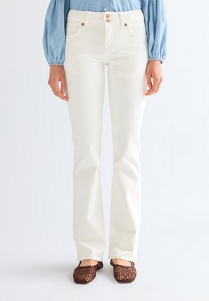 Frau trägt hoch taillierte weiße Jeans mit geradem Bein, blaues Langarmshirt und braune gewebte flache Schuhe, steht vor einem schlichten Hintergrund.