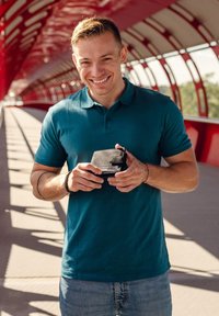 Black wallet with zipper and clasp, held by a person in a teal polo shirt. Red structure in background, creating striped shadows on the ground.