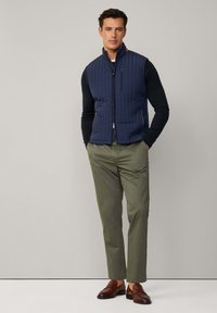 Gilet matelassé bleu marine avec col montant, fermeture éclair sur le devant et poches latérales, associé à une chemise noire à manches longues et un pantalon vert olive. Chaussures brunes.