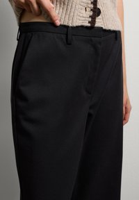 Zwarte tailleurbroek met een gladde textuur, voorzien van een platte voorkant, riemlussen en een middelhoge taille. Close-up van het taillegebied.