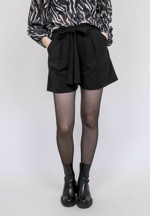 Femme portant un short taille haute noir avec une ceinture nouée, des collants noirs transparents, des bottines noires et un chemisier à manches longues à motifs.