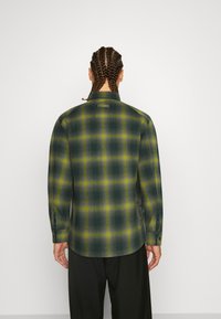 Grön och gul rutig flanellskjorta med krage, knappmanchetter och rak skärning, försedd med en liten logotyp-patch på baksidan.