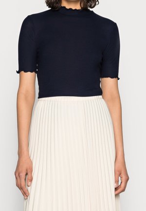 Femme portant un haut noir ajusté à manches courtes avec des bords festonnés et une jupe plissée blanche taille haute.