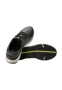 Baskets noires avec un dessus texturé, bout rond, système de laçage et une semelle intermédiaire blanche. La semelle extérieure présente un design à motifs avec une ligne d'accent jaune.