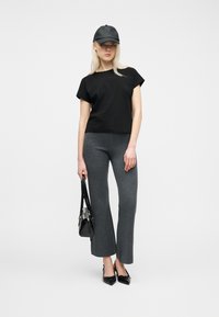 Γυναίκα που φοράει μαύρο καπέλο, μαύρο κοντομάνικο πουκάμισο, γκρι παντελόνι καμπάνα, μαύρα μυτερά παπούτσια, κρατώντας μαύρη τσάντα χειρός σε λευκό φόντο.