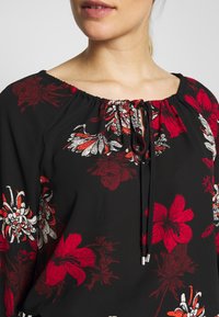 Blusa negra con estampado floral rojo y blanco, con escote fruncido y cierre de cordón con terminales de metal. Textura suave.