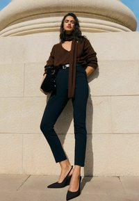 Ruskea neulepaita, jossa V-aukko ja siihen sopiva huivi, yhdistetty mustiin kapealahkeisiin housuihin ja mustiin slingback-korkokenkiin. Sileät tekstuurit.