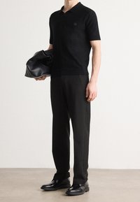 Zwarte gebreide polo met korte mouwen, met een zakdetail; gecombineerd met zwarte maatwerkbroek en glanzende zwarte schoenen. Houdt een zwarte tas vast.