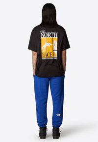 Sort bomuldstrøje med hvide grafikker på ryggen, der viser en klatrer og bjerge, kombineret med blå sweatpants og sorte støvler.