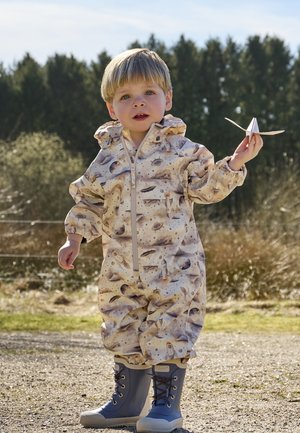 Jeune enfant en plein air tenant un avion en papier, portant une combinaison à thème spatial et des bottes de pluie bleues par une journée ensoleillée.