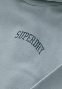 Gaiši zils treniņtērps ar mīkstu tekstūru. Priekšpusē tumši zaļā krāsā ir izdrukāts "SUPERDRY" logo izliekta fonta burtiem.
