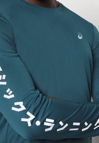 Langärmliges Sporthemd aus dunkeltürkisfarbenem Stoff, mit weißem Text entlang der Ärmel und einem kleinen Logo auf der Brust. Glatte Textur.