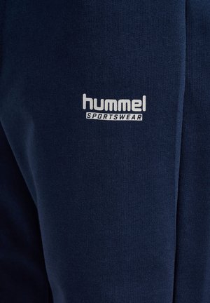 Marineblå sportbukser lavet af blødt stof med en hvid "hummel sportswear"-logo på nederste venstre side. Enkel design uden mønstre.