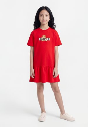 Κορίτσι όρθιο, φορώντας κόκκινο φόρεμα με κοντά μανίκια που έχει ένα αρκουδάκι και λογότυπο με κείμενο στο στήθος, συνδυασμένο με ανοιχτό ροζ αθλητικά παπούτσια.