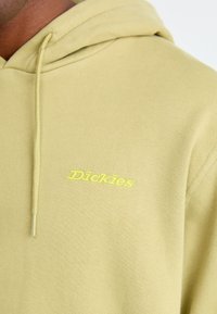 Primo piano di una felpa beige con il logo giallo "Dickies" ricamato sul petto, che mostra il cordoncino e la trama del tessuto.