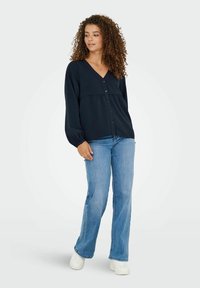 Navy blue, button-up blouse with a v-neck, puff sleeves, and a tiered design, worn with light blue flared jeans and white sneakers.