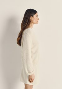 Bílý, nadýchaný cardigan z měkké tkaniny, s výstřihem do V a žebrovanými manžetami, v kombinaci s odpovídající měkkou sukní v krémové barvě.