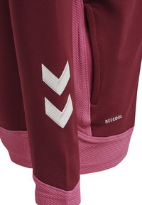 Sudadera granate con acentos en rosa. Presenta un dobladillo inferior texturizado, un bolsillo lateral y un logo de cheurón blanco en la manga. Tejido suave.