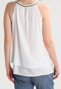 Blouse blanche sans manches avec un col haut et une découpe décorative. Présente un design superposé avec un tissu léger et doux.
