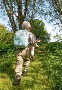 Mochila infantil azul claro con diseño de elefante y nubes, llevada por un niño con pantalones beige y zapatos marrones, caminando por un sendero de hierba.