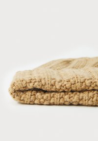 Gebreide beige muts met een gestructureerd ribpatroon, gemaakt van een zachte wolachtige stof en een omgeslagen rand voor extra warmte en comfort.