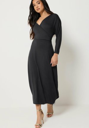 Femme portant une robe portefeuille noire à manches longues et des sandales dorées à talons hauts, debout devant un fond clair uni.