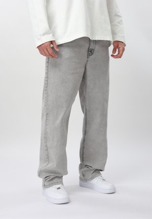 Person iført lysegrå løse jeans, hvid langærmet skjorte og hvide sneakers, der står mod en ensartet lys baggrund.