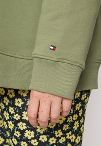 Olivgrüner Sweatshirt mit gerippten Bündchen, mit einem kleinen rot-weißen-blauen Logo am Ärmel. Darunter gelb gedruckte Hosen mit Blumenmuster.