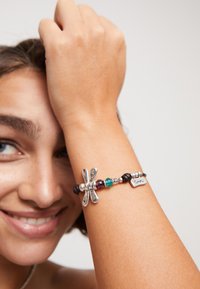 Bracelet avec un charm en libellule en argent, agrémenté de perles colorées, d'accents en cuir et d'une étiquette en métal. Il présente des textures variées et un mélange de matériaux.