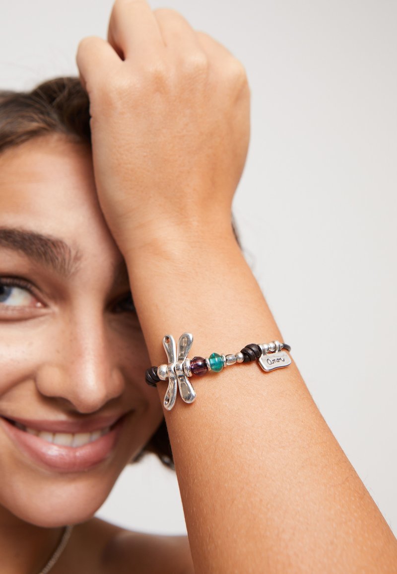 Bracelet avec un charm en libellule en argent, agrémenté de perles colorées, d'accents en cuir et d'une étiquette en métal. Il présente des textures variées et un mélange de matériaux.