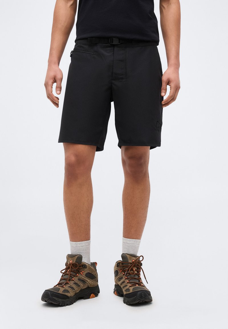 Άνδρας που φοράει μαύρο σορτς, γκρι κάλτσες και καφέ ορειβατικά παπούτσια, στέκεται μπροστά σε απλό ανοιχτό φόντο.