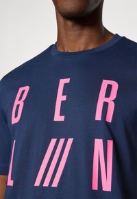 Marineblå bomulds T-shirt med iøjnefaldende, pink bogstaver, der staver "BERLIN" med tre vandrette linjer. Crew neck-design med en glat tekstur.