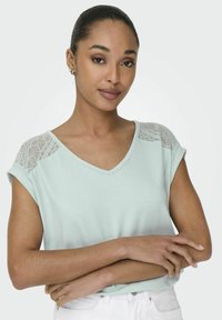 Jasnozielona koszulka z dekoltem w serek, krótki rękaw, z koronkowymi detalami na ramionach, wykonana z miękkiego materiału o gładkiej teksturze.
