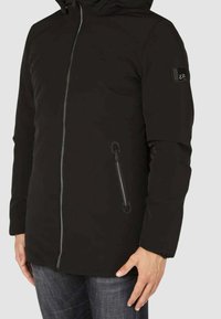Giacca nera con cappuccio, chiusura frontale con zip, polsini elasticizzati e tasche laterali. Tessuto liscio con design minimalista e patch del logo sulla manica.