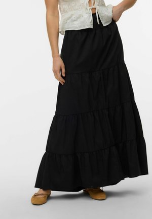 Kvinde iført et lagdelt sort maxinederdel, hvid ærmeløs øjlet-top og brune balletflade sko, står med den ene hånd på hoften.