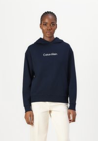 Námornícky modrá mikina s kapucňou vyrobená z mäkkej látky, s bielym logom "Calvin Klein" na hrudi a so žebrovanými manžetami a spodným okrajom.