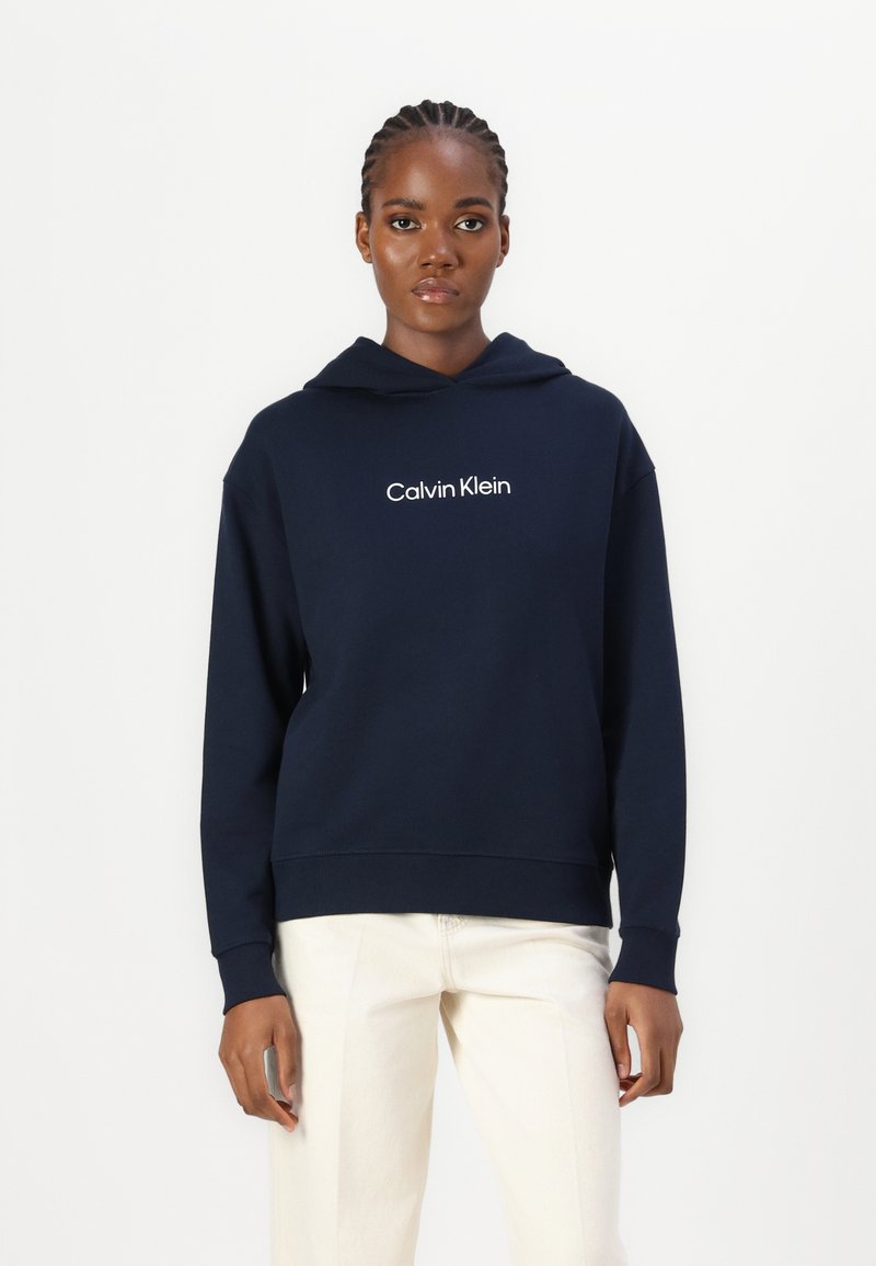 Námornícky modrá mikina s kapucňou vyrobená z mäkkej látky, s bielym logom "Calvin Klein" na hrudi a so žebrovanými manžetami a spodným okrajom.
