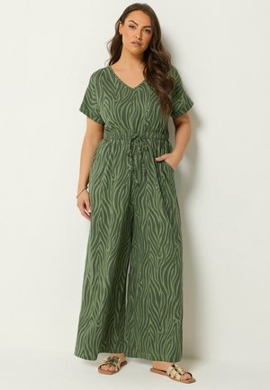 Kobieta ubrana w zielony kombinezon w zebra print, z dekoltem w serek, ściągaczem w talii i szerokimi nogawkami, w połączeniu z beżowymi sandałami wsuwanymi.