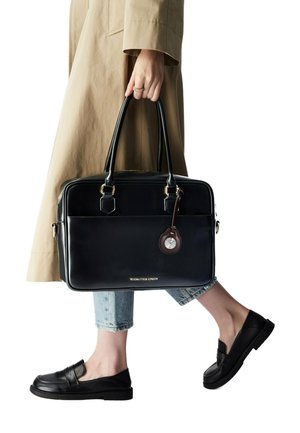 Sac à main en cuir noir au design structuré avec deux poignées, orné d'une étiquette marron avec un détail d'horloge. Porté avec un jean et des chaussures slipes noires.