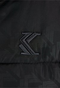 Sort stoff har et stilrent, brodert logo i en mørkere nyanse av svart. Designet inkluderer subtile geometriske mønstre for ekstra tekstur.