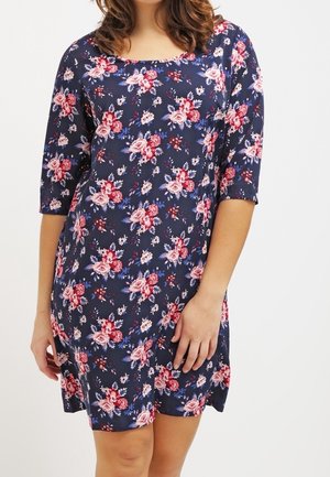 Frau in einem marineblauen knielangen Kleid mit rotem und pinkfarbenem Blumenmuster und dreiviertel Ärmeln, die vor einem einfarbigen Hintergrund steht.