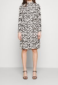 Black and white chevron-patterned dress with long sleeves, a belted waist, and a button-up front. Worn with black strappy sandals.